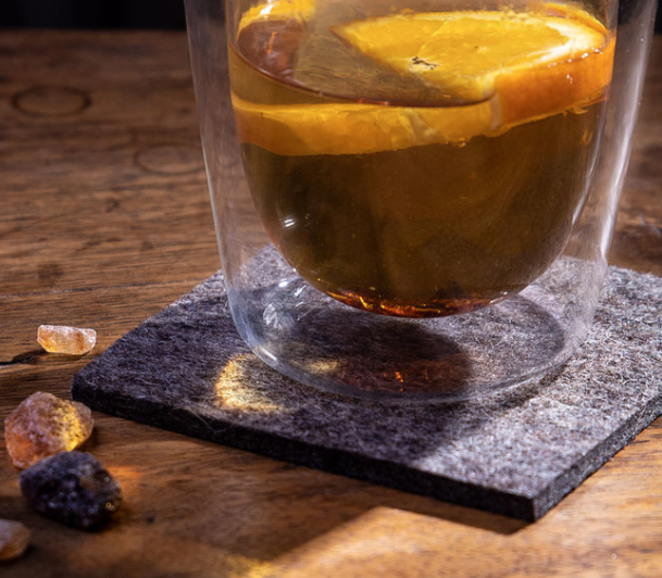  Glass of orange-infused drink on dark grey wool felt coaster on wooden surface